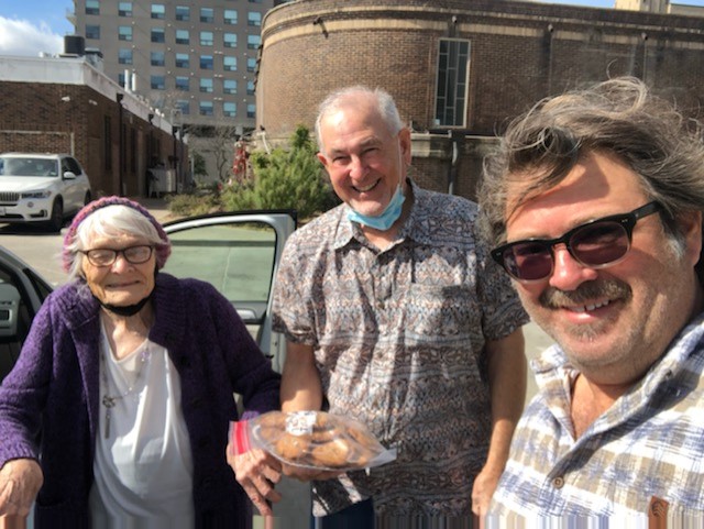 Carolyn "Bertrand" Hodges, David Behrman, and Mitch Cohen stand together outdoors, smiling warmly after celebrating Carolyn’s 95th birthday. On the left, Carolyn, an elderly woman with white hair and glasses, wears a purple cardigan and white top, holding a walker. In the center, David, with short gray hair and a patterned shirt, holds a tray of baked goods, his face mask below his chin. On the right, Mitch, with gray hair and sunglasses, wears a plaid shirt, standing near an open car door. The background features a brick building, a multi-story structure, and greenery under a clear sky.